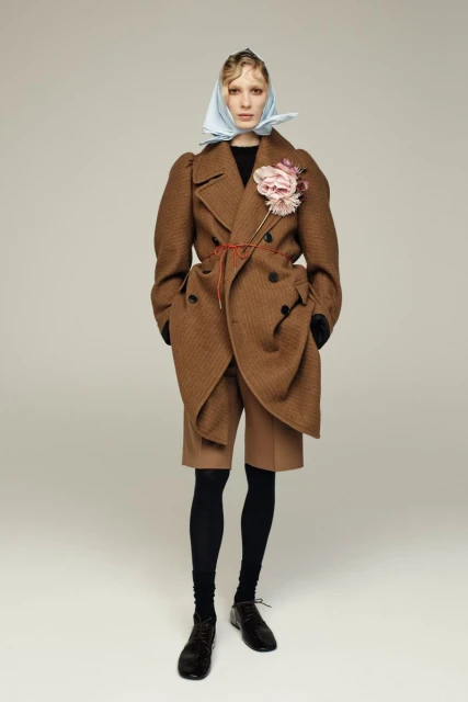 Full-shot fashion portrait of a woman wearing a brown overcoat, shorts, tights, and a headscarf in a bright studio setting.