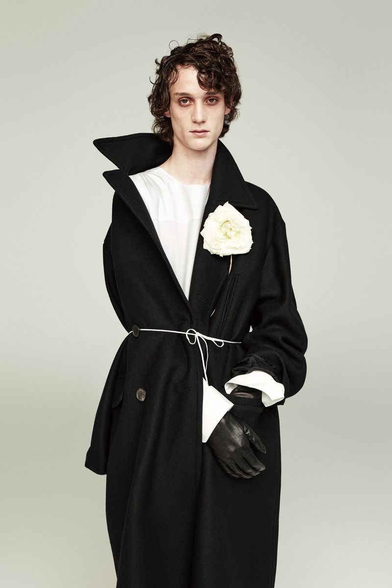 Male model in a black overcoat with a white flower on the lapel, cinched with a white string belt against a neutral backdrop.