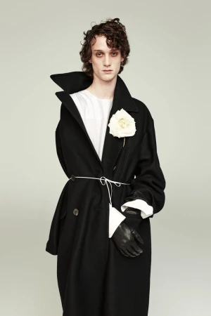 Male model in a black overcoat with a white flower on the lapel, cinched with a white string belt against a neutral backdrop.