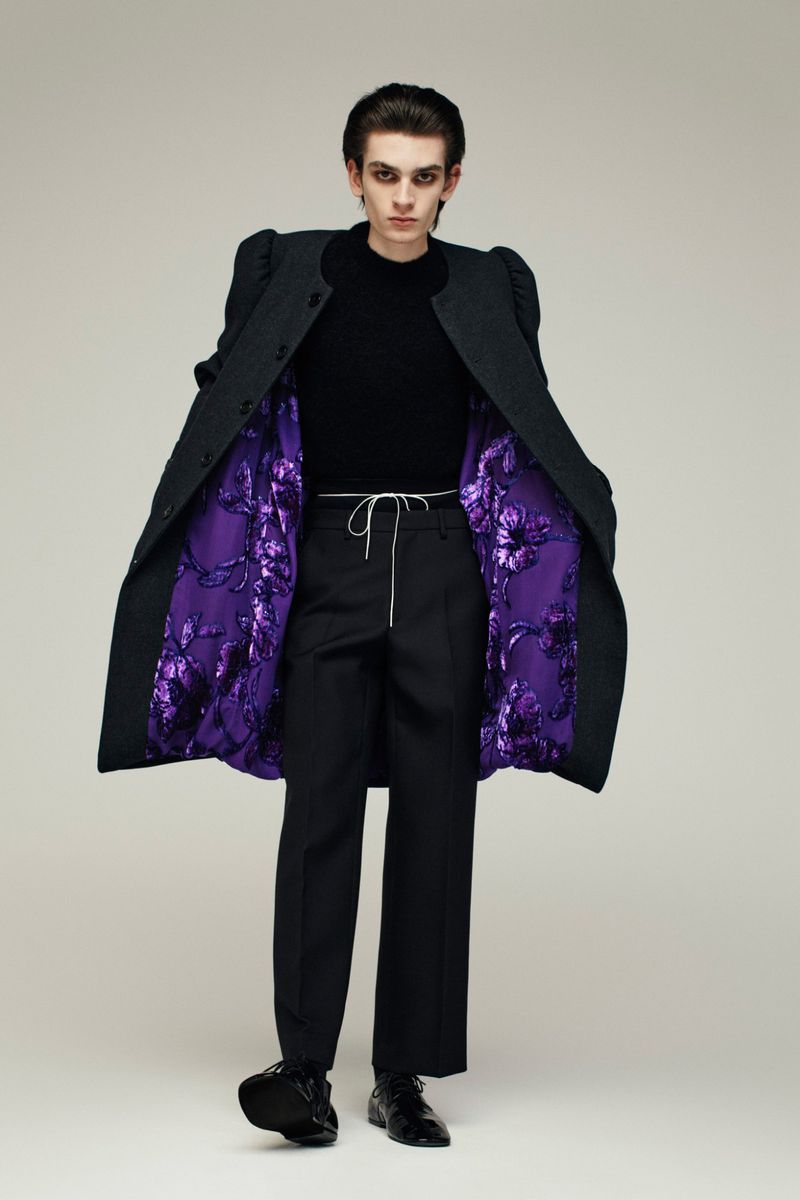 Model wearing a dark structured coat with vibrant purple floral lining over black trousers against a neutral background