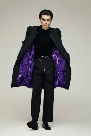 Model wearing a dark structured coat with vibrant purple floral lining over black trousers against a neutral background