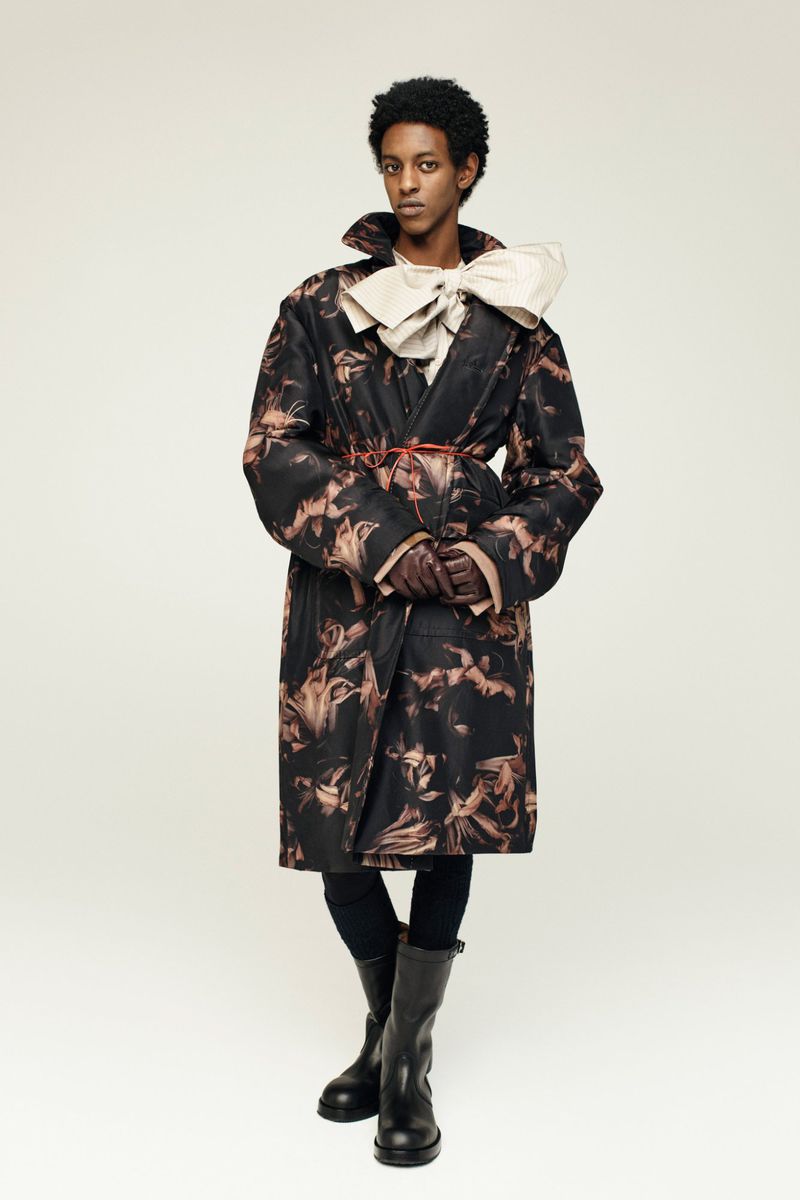 A person stands in a photo studio, wearing a patterned trench coat, black leggings, and boots, with a large cream bow at the neck.