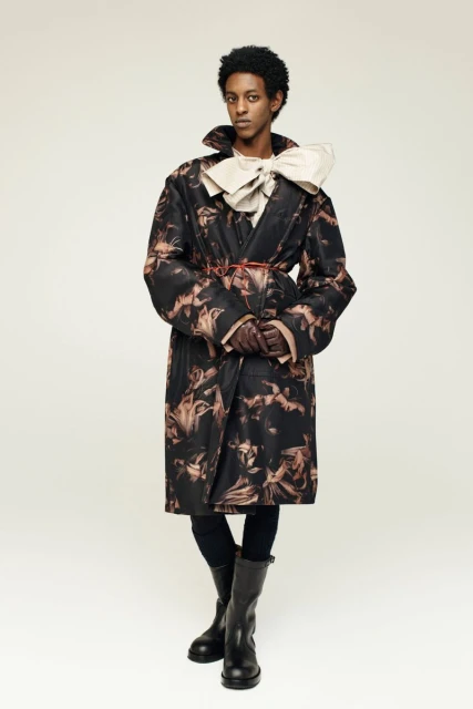A person stands in a photo studio, wearing a patterned trench coat, black leggings, and boots, with a large cream bow at the neck.