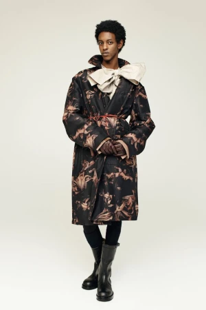 A person stands in a photo studio, wearing a patterned trench coat, black leggings, and boots, with a large cream bow at the neck.