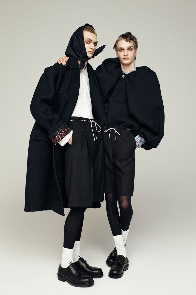 Two slender fashion models pose in coordinated black outfits against a neutral backdrop, showcasing modern androgynous style.