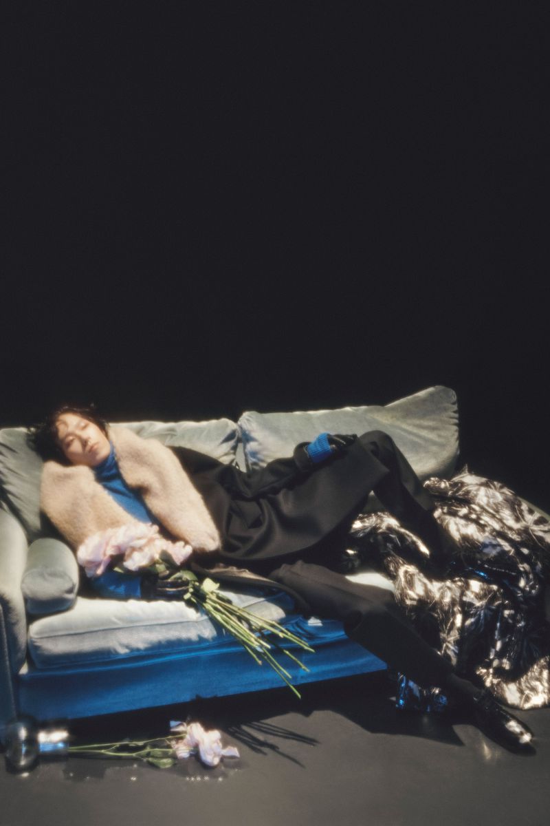 Fashion shot of a woman reclining on a teal couch with a bouquet of flowers in a studio setting against a dark background.