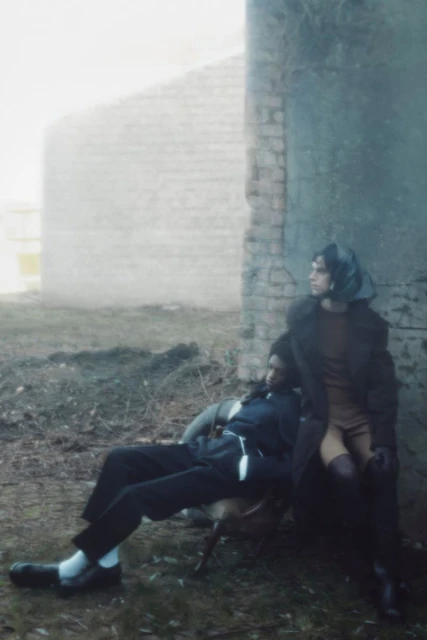Two people in vintage fashion, one seated and one leaning against a weathered building, in a muted outdoor environment with moody light.