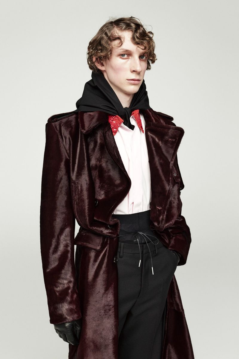 Male model in burgundy coat with black hooded sweatshirt, red patterned dress shirt, and black pants in a fashion studio shot.
