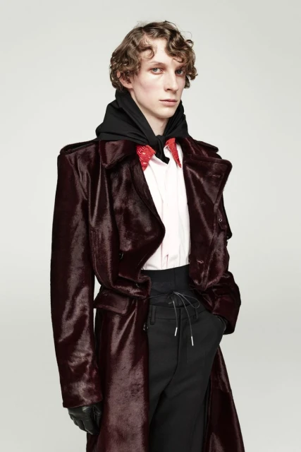 Male model in burgundy coat with black hooded sweatshirt, red patterned dress shirt, and black pants in a fashion studio shot.