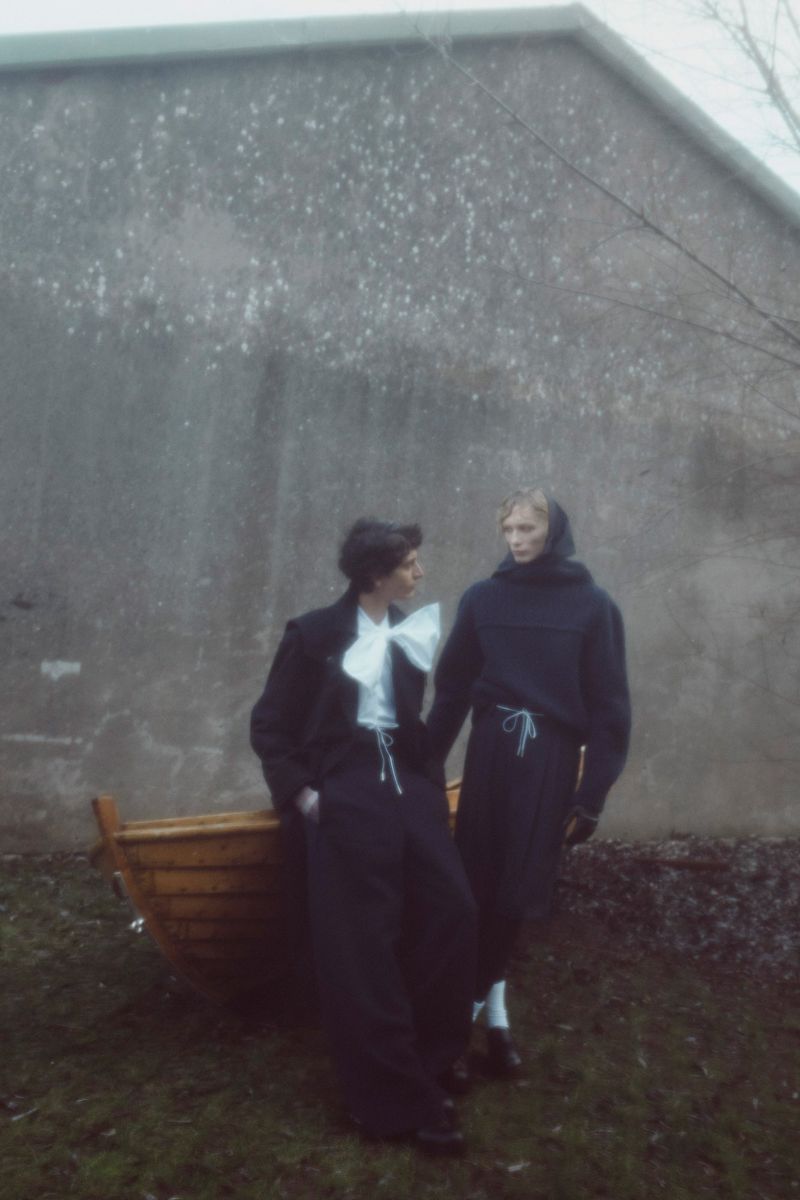 Two people stand on a grassy yard next to a wooden boat in front of a concrete building on a cloudy day. Fashion editorial.