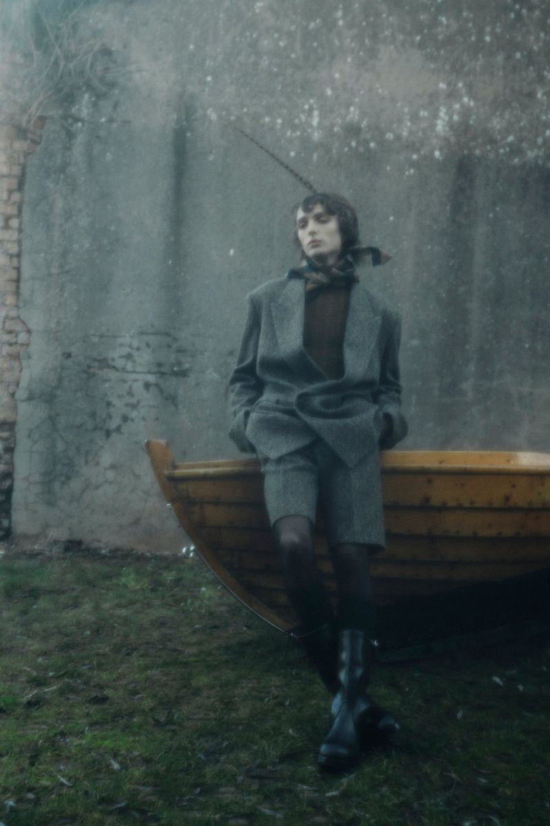 Stylish woman in a tweed suit poses by a weathered wooden boat against an old brick wall in an outdoor setting. Dark, muted tones.