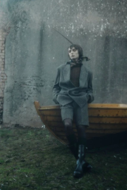 Stylish woman in a tweed suit poses by a weathered wooden boat against an old brick wall in an outdoor setting. Dark, muted tones.