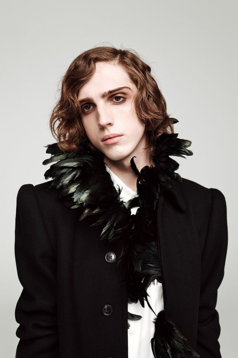 Portrait of a young man with wavy brown hair wearing a black coat adorned with black feathers against a neutral grey studio background