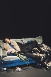 Model reclining on a blue velvet sofa holding flowers against a dark background in Dries Van Noten lookbook
