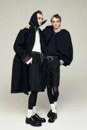 Two male models in dark coats and shorts posing together against a plain neutral background for Dries Van Noten