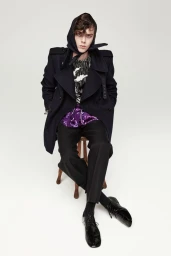 Model wearing a dark coat and headscarf seated on a small wooden stool against a white background