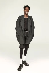 Model standing against a seamless white background wearing an oversized grey blazer, dark shorts, white socks, and black shoes