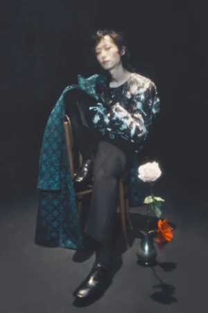 Model seated on a wooden chair holding a patterned teal coat next to a rose in a vase against a dark backdrop