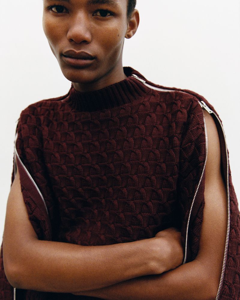 Studio shot of a stylish adult male wearing a patterned knit sweater with zipper details and arms crossed against a neutral background.