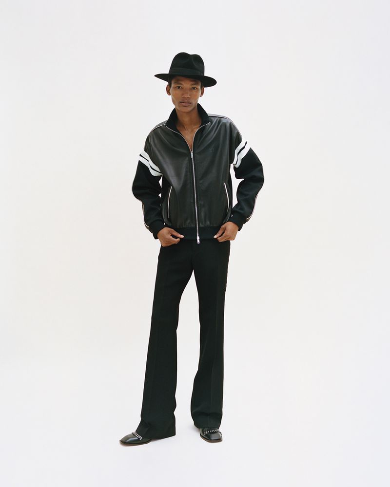 Full-length shot of an adult male model in a black track jacket with white stripes, black bell-bottoms, and black hat against a white backdrop.