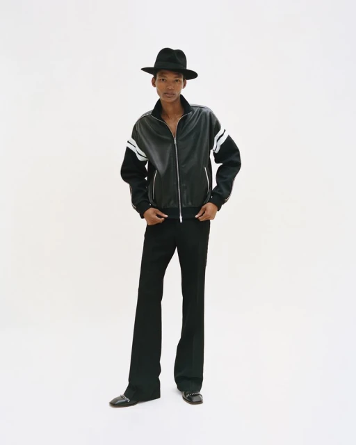Full-length shot of an adult male model in a black track jacket with white stripes, black bell-bottoms, and black hat against a white backdrop.