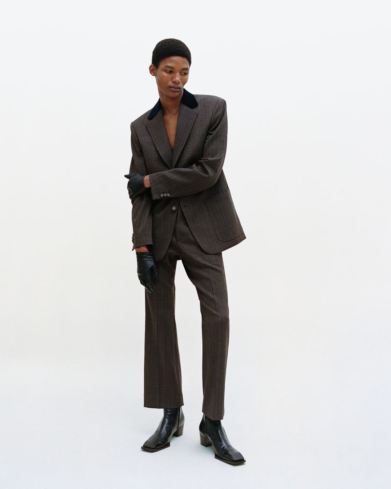 A slender, dark-skinned adult fashion model in a pinstripe suit poses in a photo studio with a plain white backdrop for a fashion editorial.