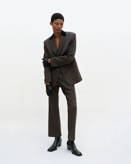 A slender, dark-skinned adult fashion model in a pinstripe suit poses in a photo studio with a plain white backdrop for a fashion editorial.
