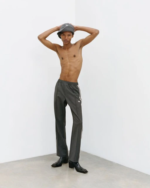 A slender shirtless man with a hat poses in gray pants and black shoes against a simple white corner backdrop, creating a minimalist fashion shot.