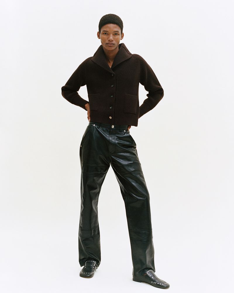 Full shot of a person in a dark brown cardigan sweater and black leather-look trousers against a white backdrop in a studio.