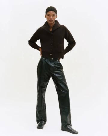 Full shot of a person in a dark brown cardigan sweater and black leather-look trousers against a white backdrop in a studio.