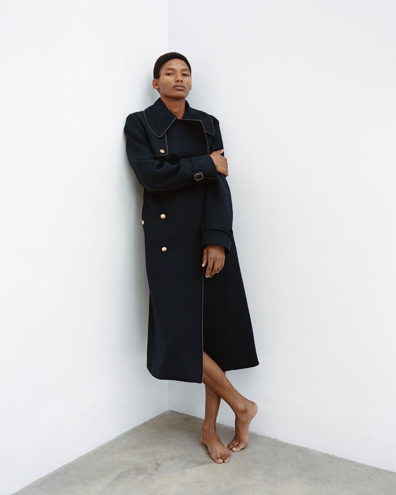 An adult woman poses indoors wearing a long navy coat with gold buttons. She is barefoot, leaning against a corner in a casual pose.