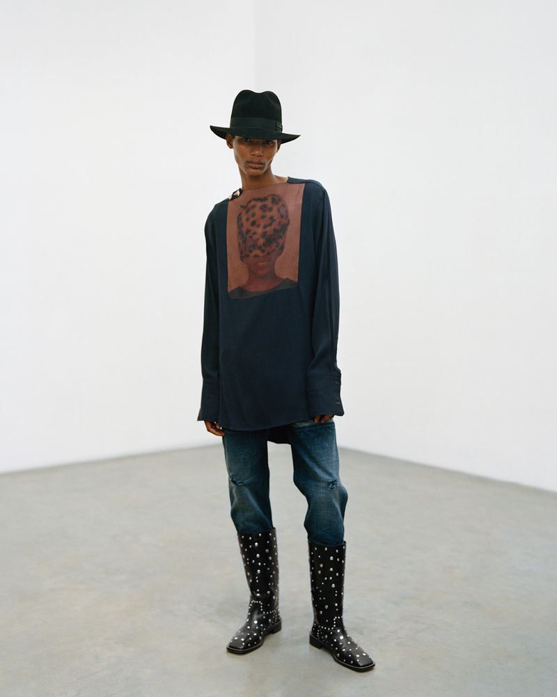 Full-length portrait of a person in a hat, long-sleeve shirt with a graphic, jeans, and polka-dot boots, posing in a white corner studio.