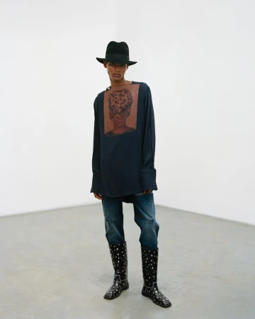 Full-length portrait of a person in a hat, long-sleeve shirt with a graphic, jeans, and polka-dot boots, posing in a white corner studio.