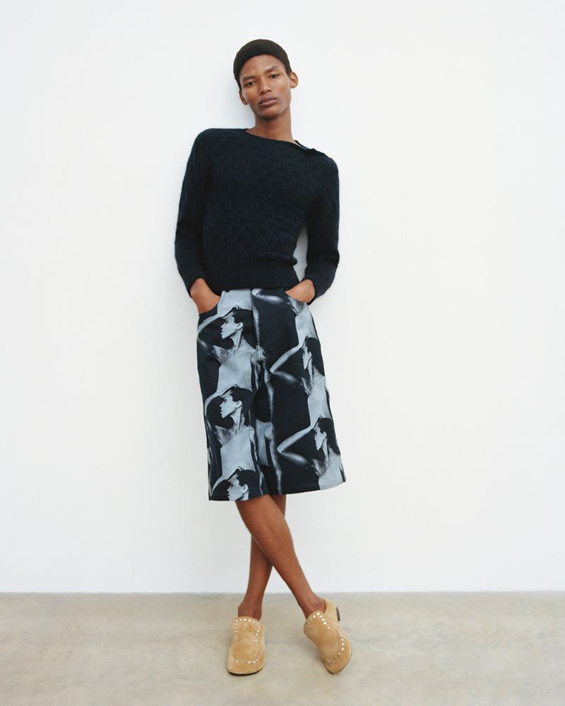 Full-body studio shot of a young adult man in a dark sweater, patterned culottes, and heeled mules standing against a white wall.