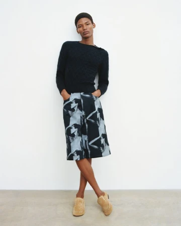 Full-body studio shot of a young adult man in a dark sweater, patterned culottes, and heeled mules standing against a white wall.