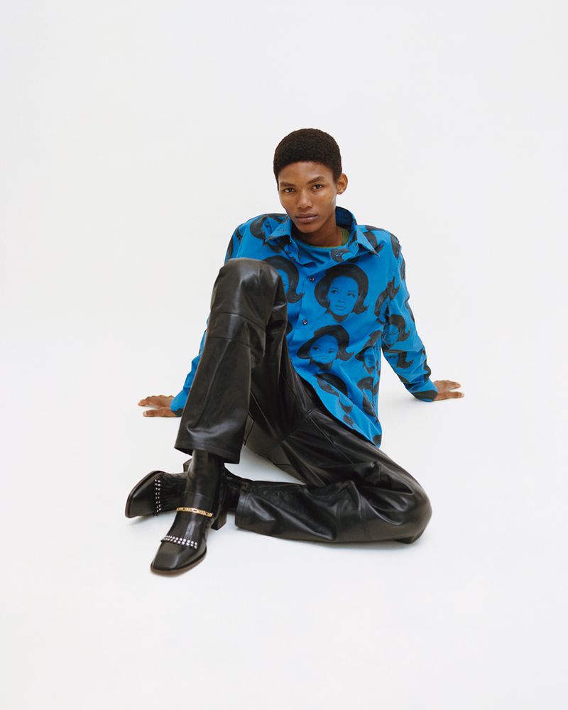 A person with short hair sits in a white studio wearing a blue printed button-down shirt, black leather pants and boots with silver chain detail.