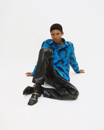 A person with short hair sits in a white studio wearing a blue printed button-down shirt, black leather pants and boots with silver chain detail.