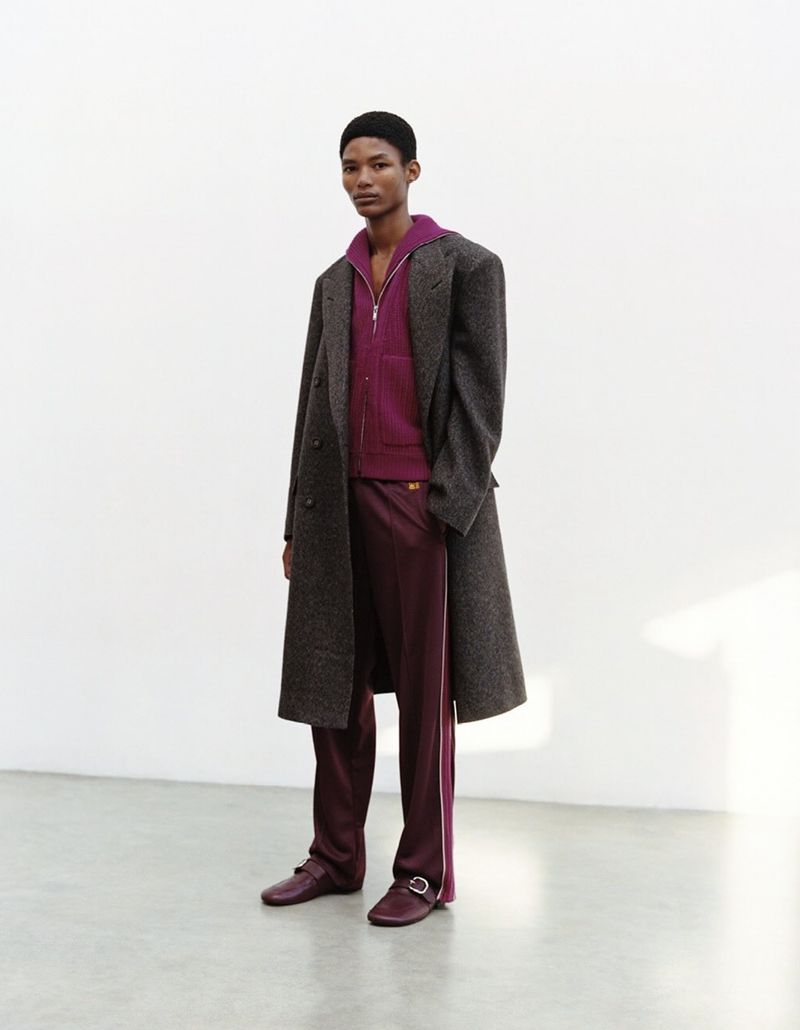 A dark-skinned male model poses in a photo studio wearing a gray overcoat, ribbed purple jacket, satin maroon pants, and matching dress shoes.
