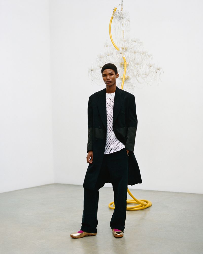 An African American adult male stands in a modern art gallery wearing a black coat, white top, wide-legged trousers and gold loafers.