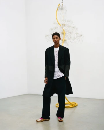 An African American adult male stands in a modern art gallery wearing a black coat, white top, wide-legged trousers and gold loafers.