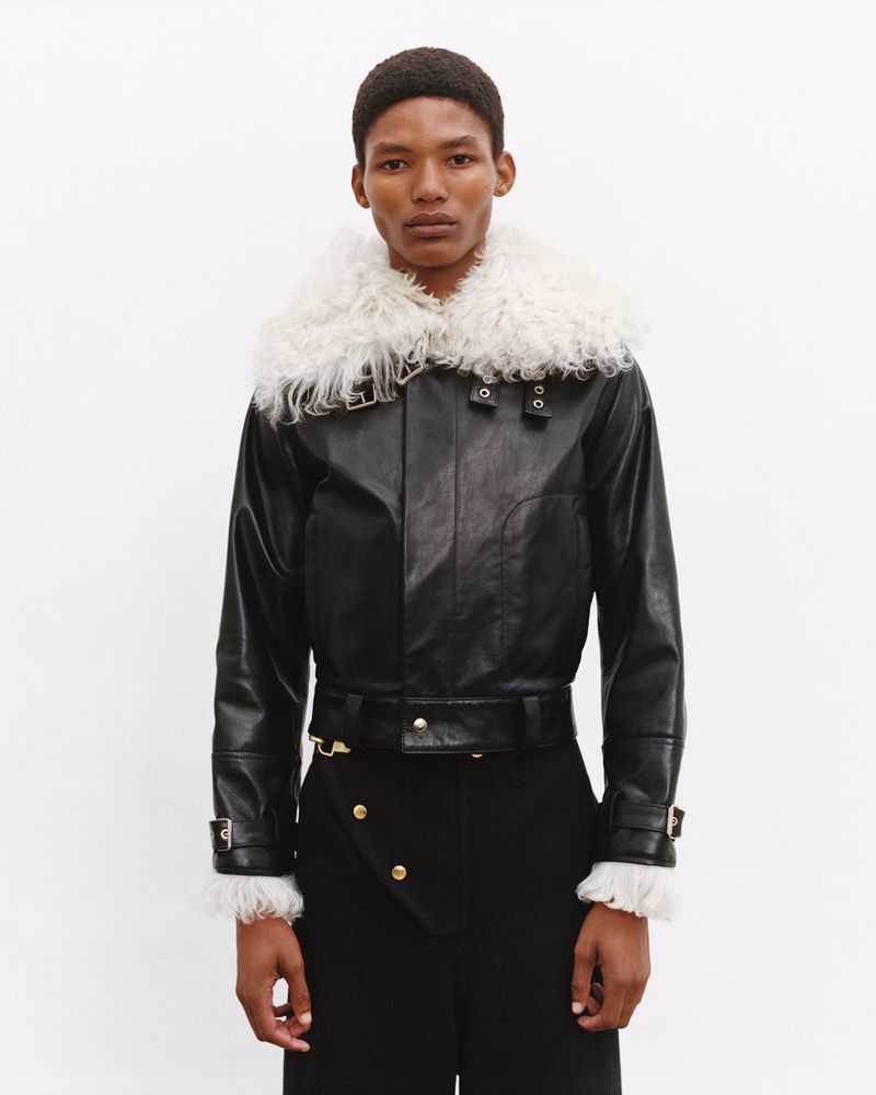 Dark-skinned male model wearing a black leather jacket with a white fur collar and black dress pants, posing against a white backdrop.