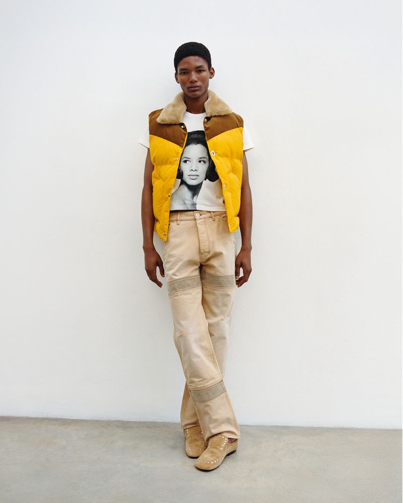 Full-length shot of an adult male wearing a yellow vest, graphic t-shirt, and beige trousers, posing against a white studio backdrop.