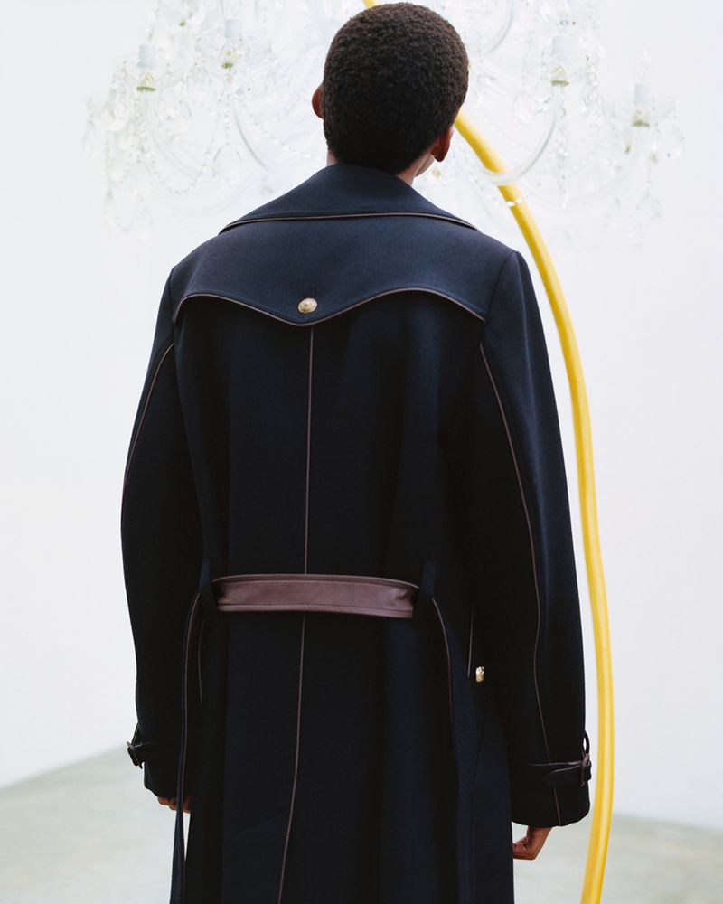 Back view of a dark-haired person wearing a navy belted trench coat indoors, standing near a yellow sculpture and transparent chandelier.