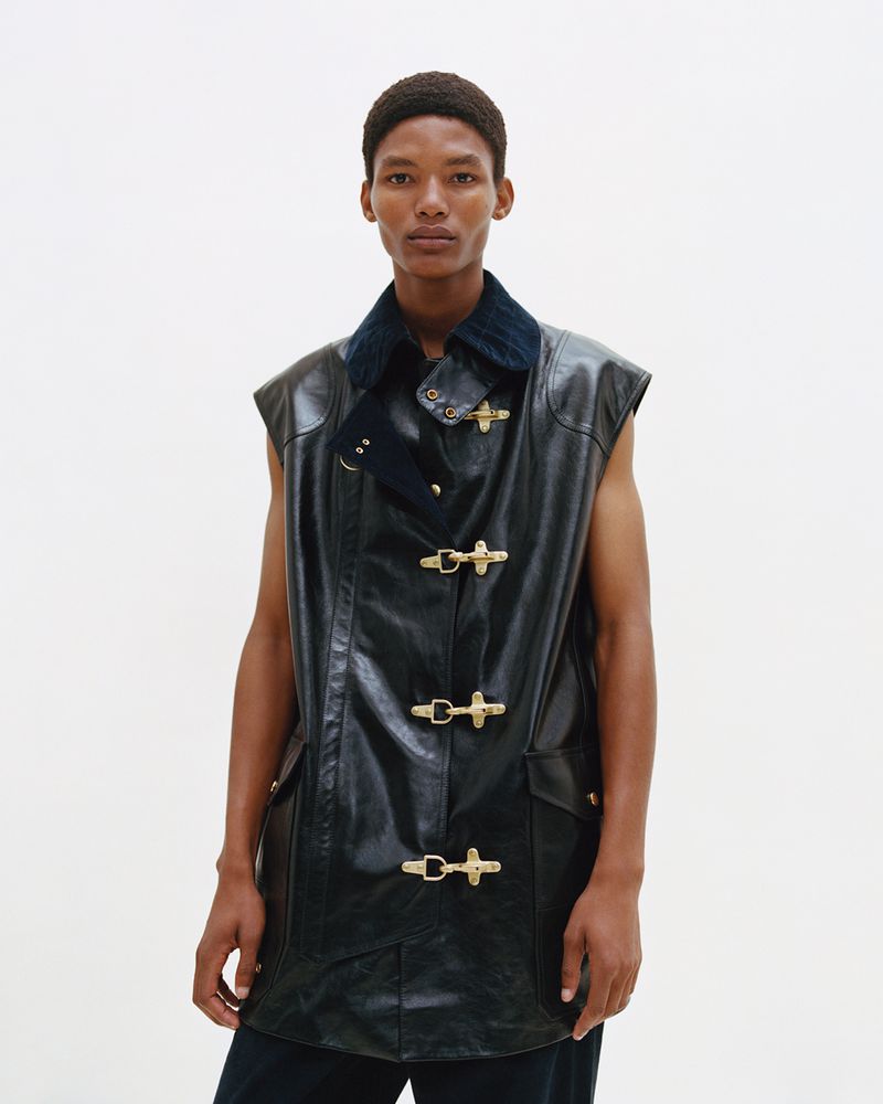A man poses in a sleeveless black leather jacket with metal clasps against a white backdrop in a fashion studio shot.