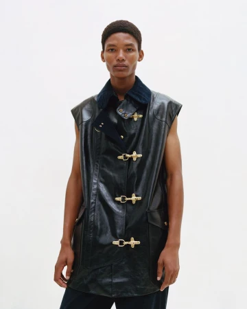 A man poses in a sleeveless black leather jacket with metal clasps against a white backdrop in a fashion studio shot.