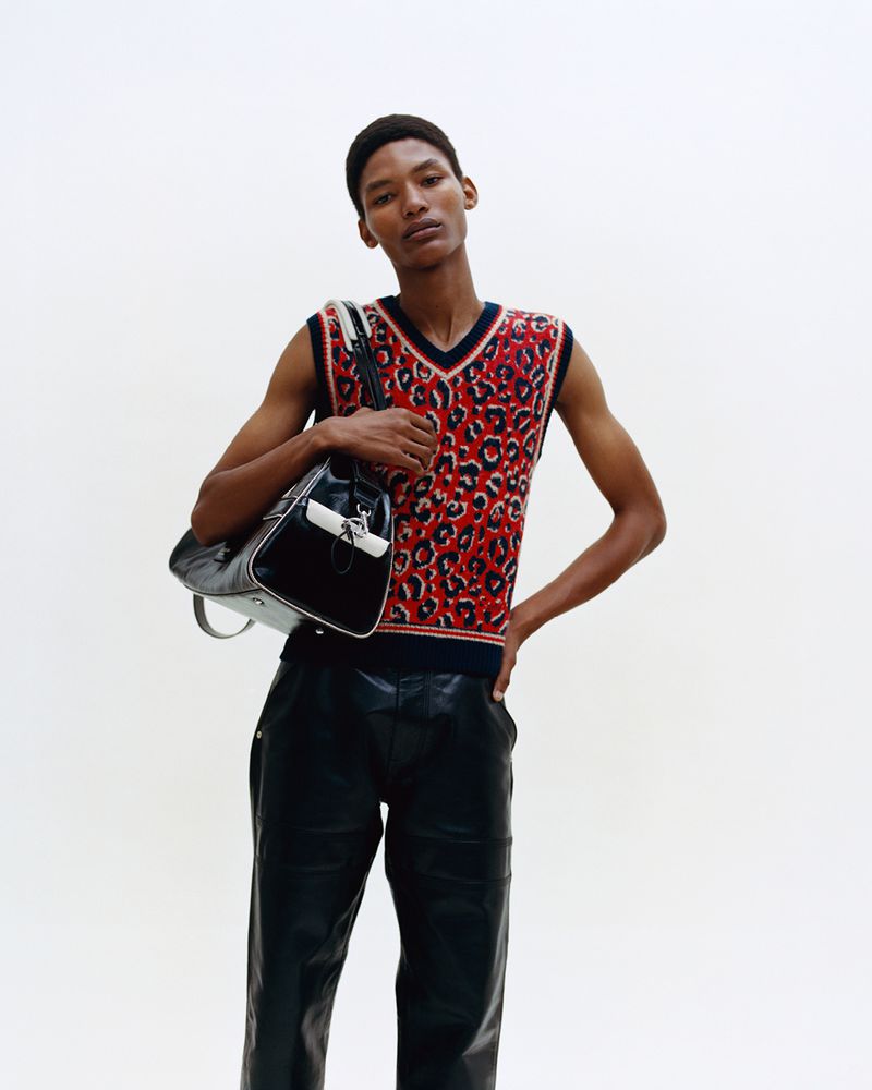 Adult male poses in a sleeveless red leopard print vest and black leather pants, holding a duffel bag against a plain white backdrop.