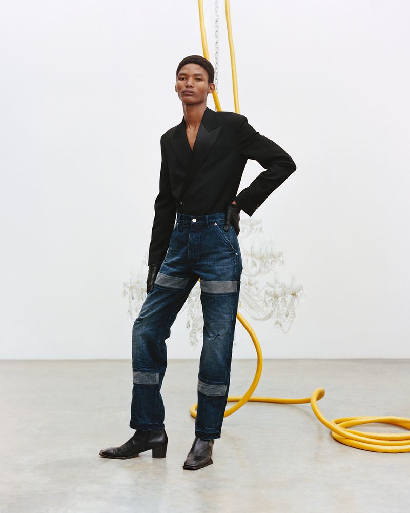 A fashion shot of an androgynous model wearing a black blazer and dark blue jeans, standing in a studio against a white wall with a yellow pipe.