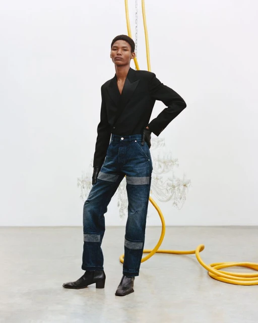 A fashion shot of an androgynous model wearing a black blazer and dark blue jeans, standing in a studio against a white wall with a yellow pipe.
