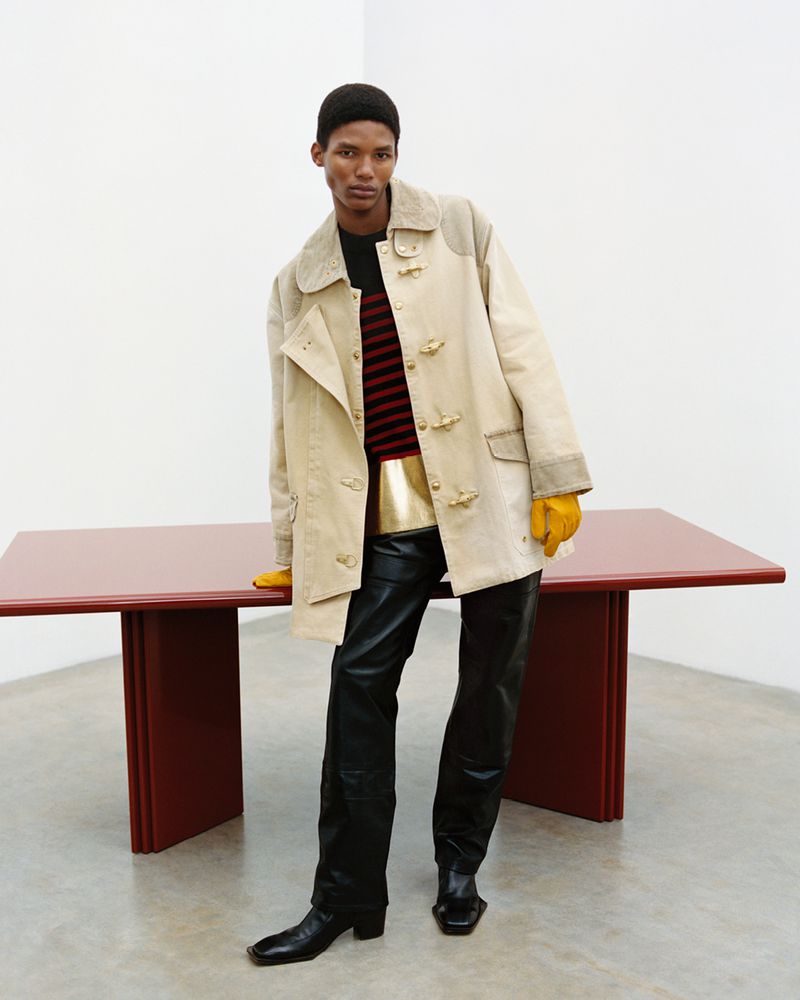 Stylish adult male in beige coat, black leather pants, and boots stands next to a red table against a plain white background.