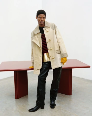 Stylish adult male in beige coat, black leather pants, and boots stands next to a red table against a plain white background.